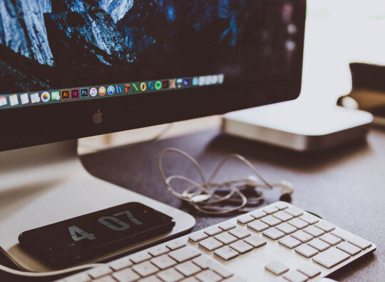 Close-up of a desk with an iMac and a smartphone displaying the time